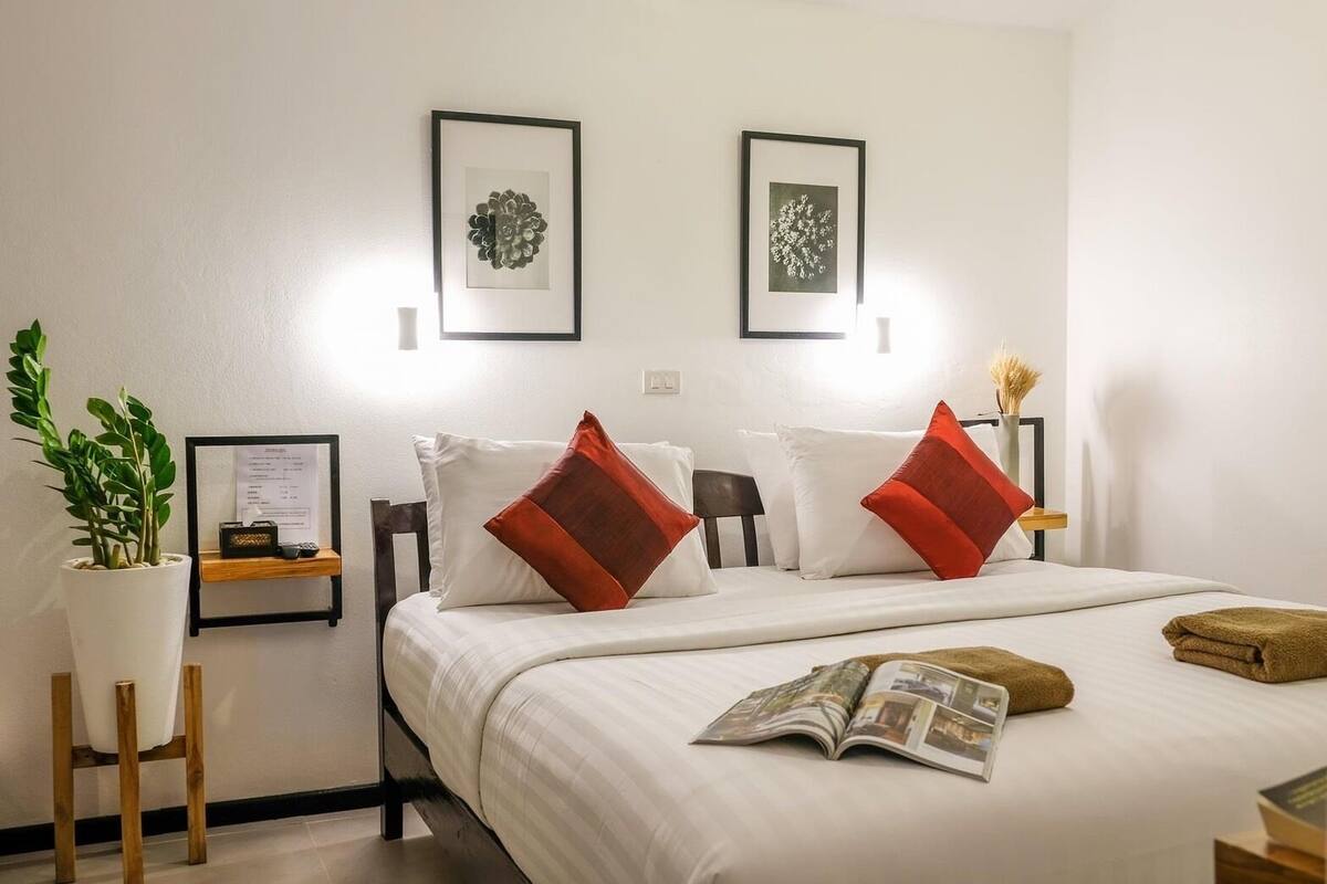 A neatly arranged hotel room with a bed, pillows, a bedside table with a lamp, a framed picture on the wall, and a potted plant.