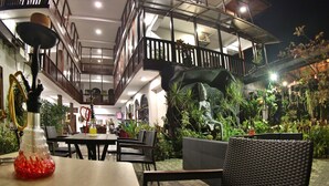 An outdoor dining area with multiple tables and chairs, surrounded by lush greenery and a decorative rock formation. - Hotel Sunray (Kandy)