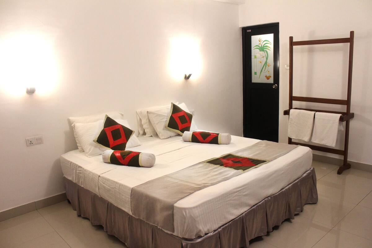 A neatly made bed with a brown skirt, two wall-mounted lights, a black door with a floral design, and a wooden towel rack.