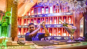 A bartender preparing a drink at a bar with shelves of liquor bottles and hanging glasses. - Hotel Sunray (Kandy)