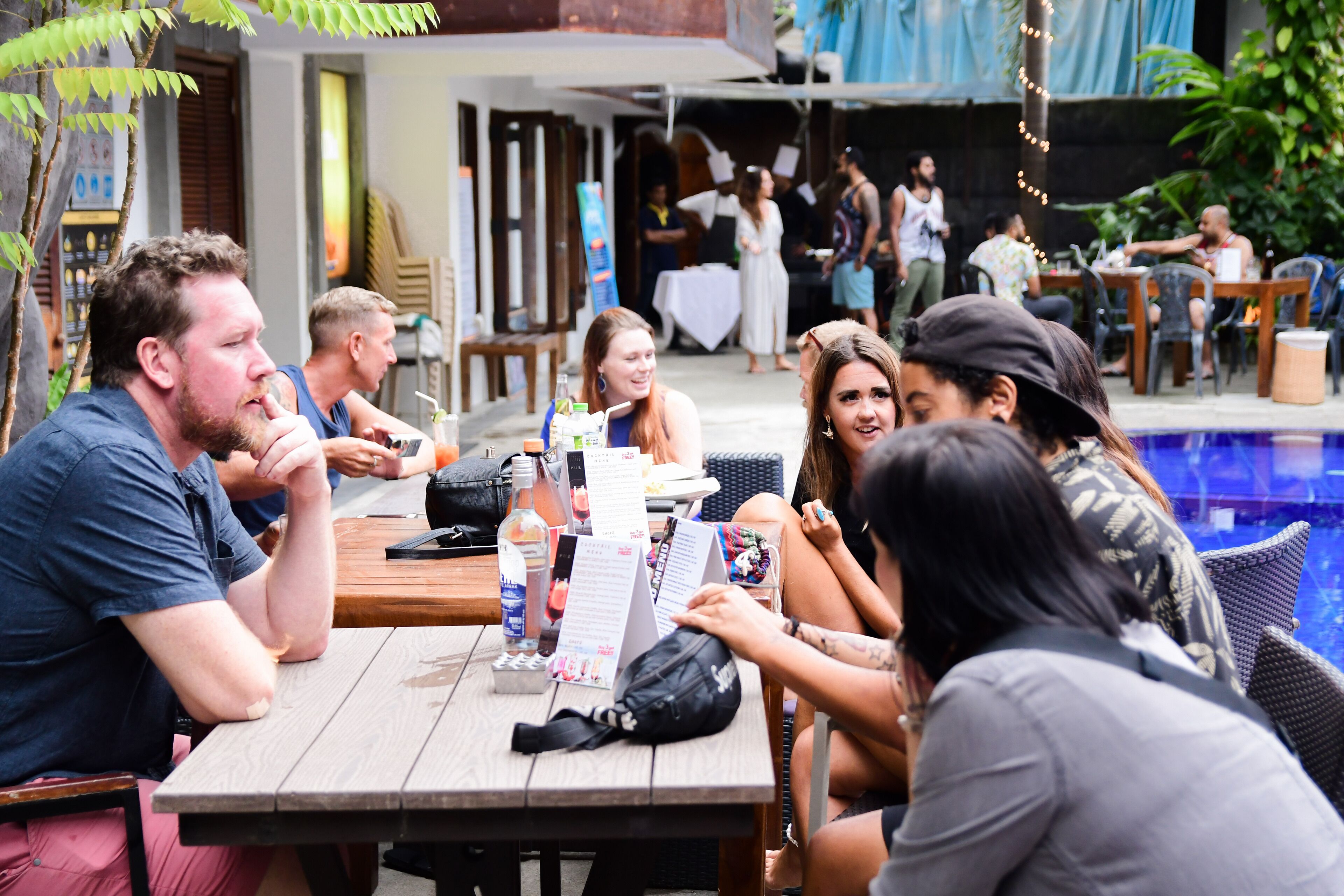 A group of people seated at an outdoor table with a menu, surrounded by a pool and greenery.