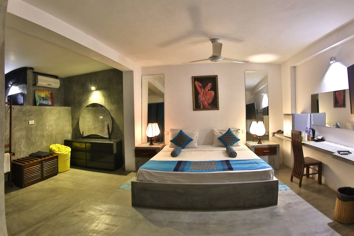 A hotel room with a large bed, a desk, a chair, a mirror, a ceiling fan, and a bathroom with a shower and sink.