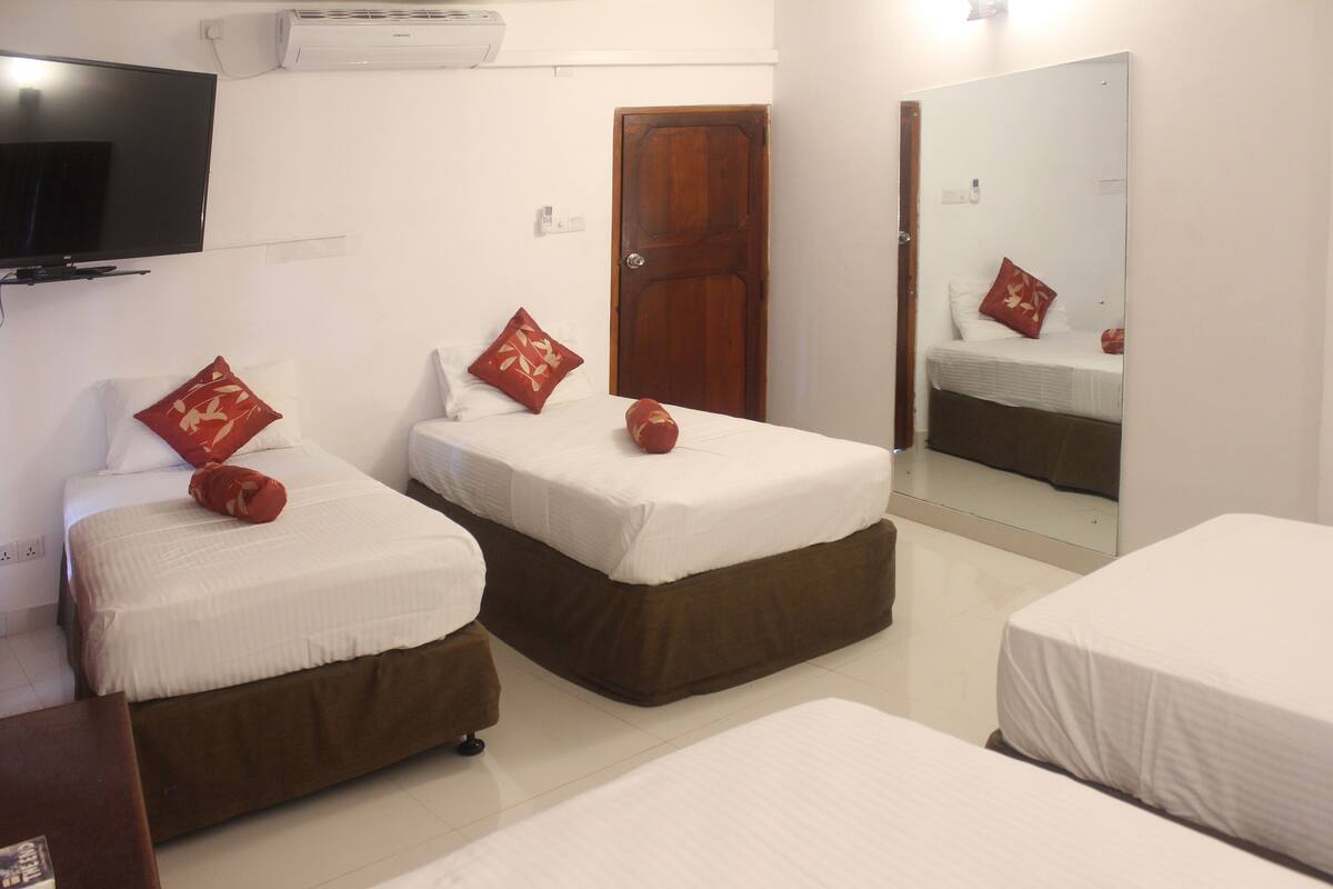 A hotel room with two single beds, each with a brown headboard, white bedding, and red decorative pillows. A flat-screen TV is mounted on the wall above the left bed.