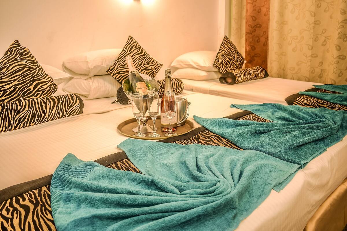 A hotel room with two beds, zebra-patterned pillows, a tray with champagne bottles and glasses, and teal towels.