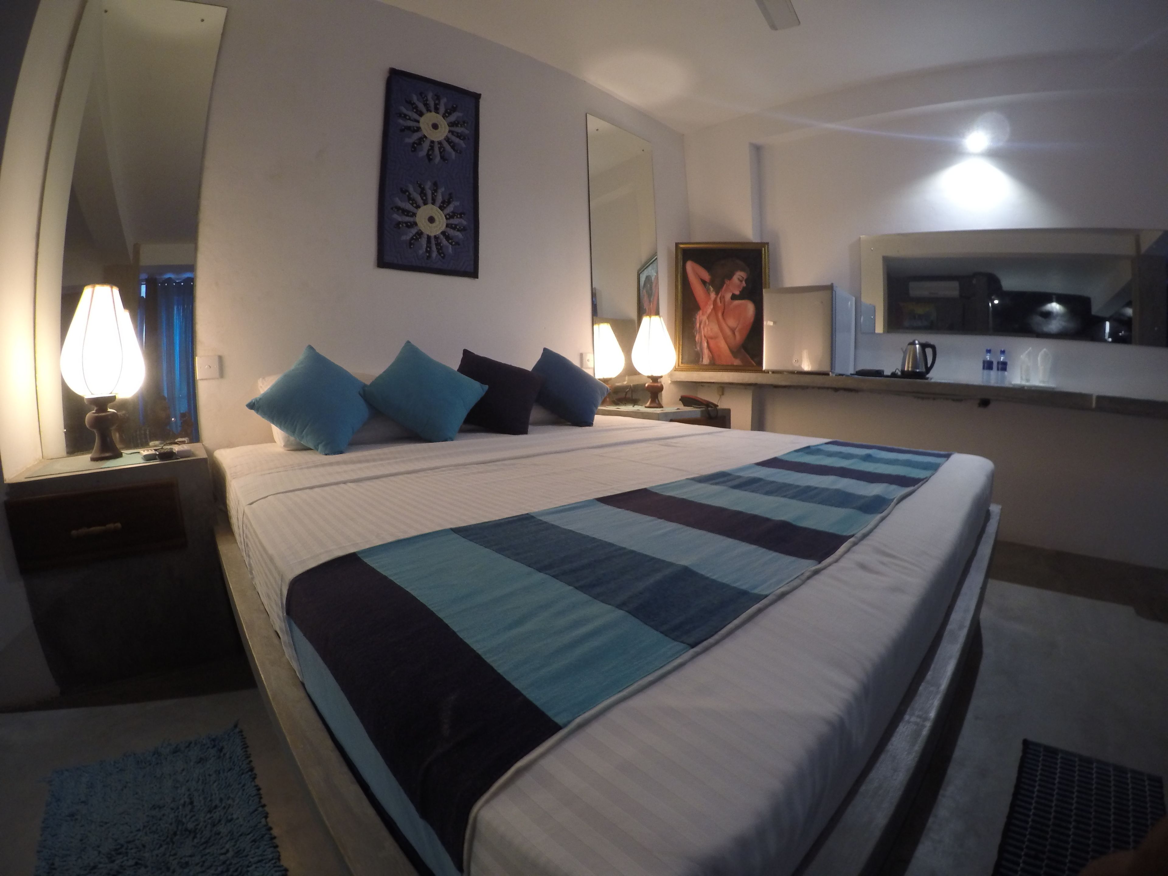 A hotel room with a large bed, a desk, a mirror, a painting, and a kitchenette.
