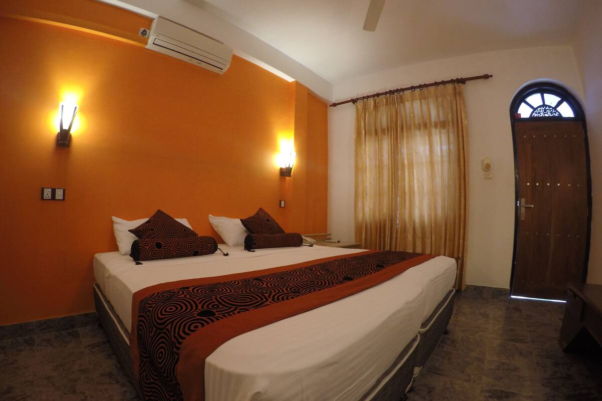 A hotel room with a large bed, two wall-mounted lights, an air conditioner, a wooden door, and a window with curtains.