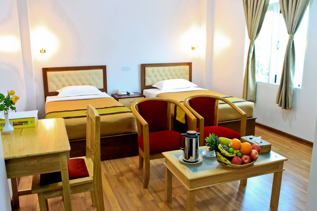 Ein Hotelzimmer mit zwei Betten, einem Holztisch mit Obstschale, einem roten Sessel und einem kleinen Tisch mit weißer Vase.