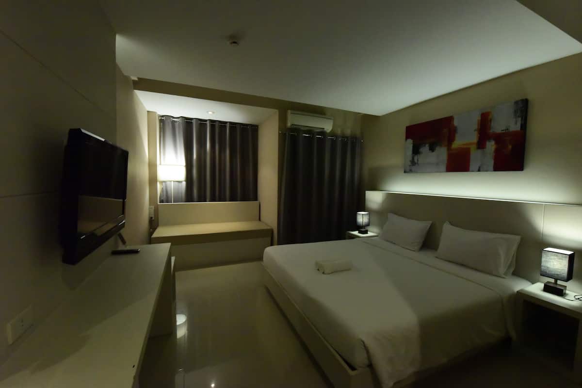 Habitación de hotel con cama, mesitas de noche, televisor, ventana con cortinas y un cuadro en la pared.