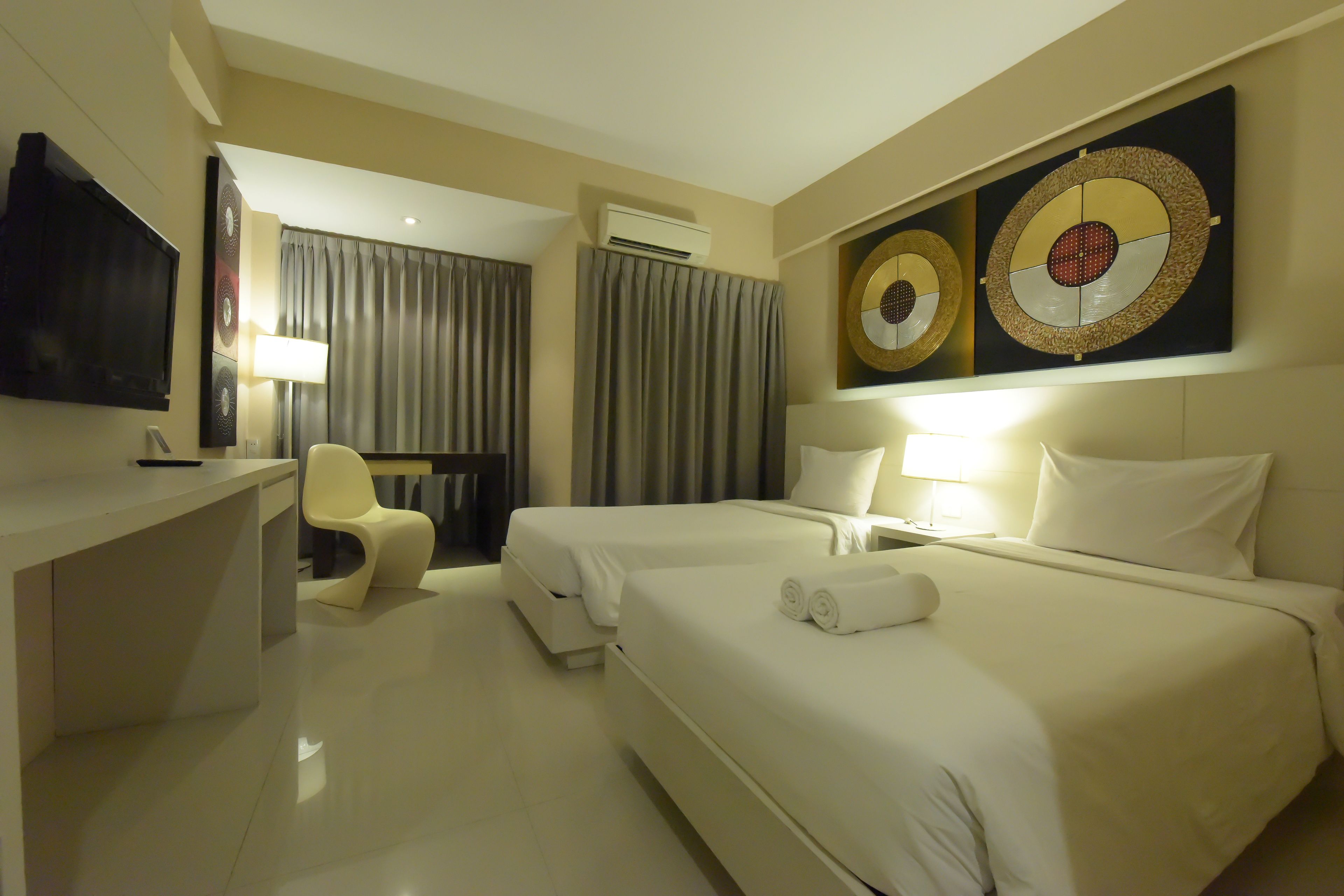 A hotel room with two beds, a desk, a chair, a TV, and artwork on the wall.