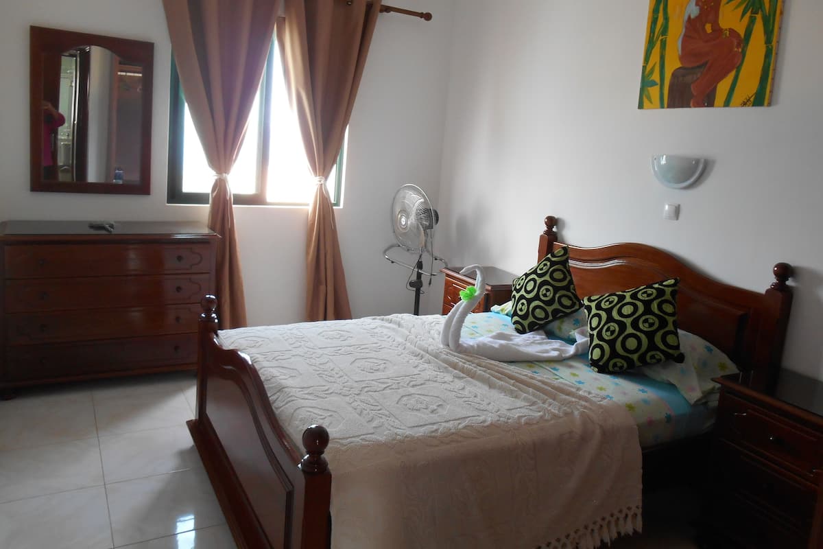 A bedroom with a wooden bed, a dresser, a bedside table, a fan, and a painting on the wall.