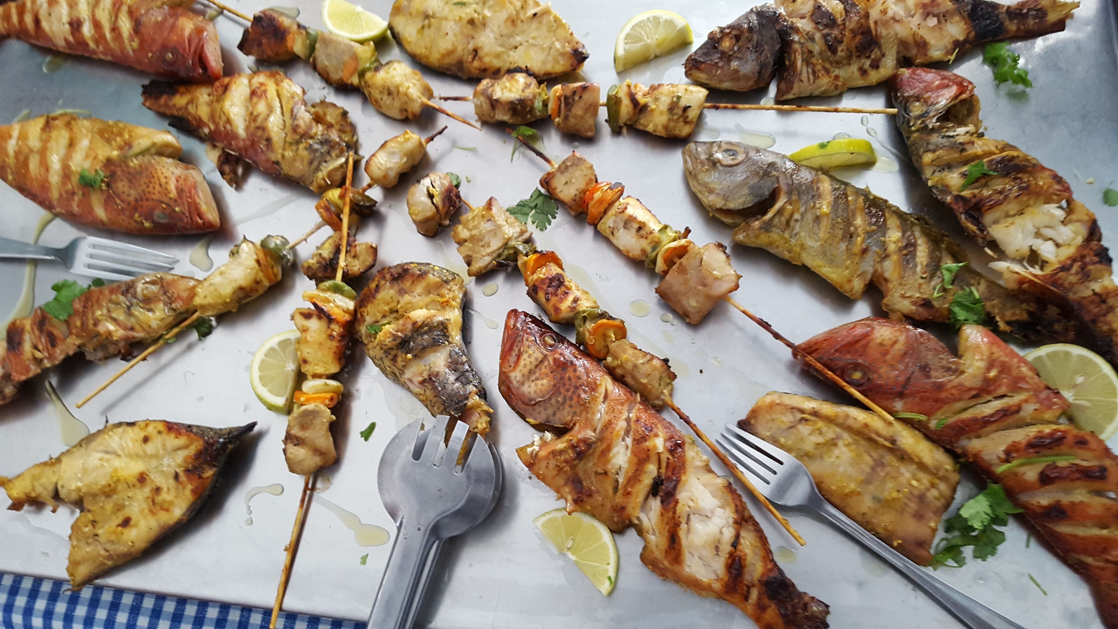 Diversos espetos de marisco grelhado e peixe numa travessa, decorados com fatias de limão e salsa.