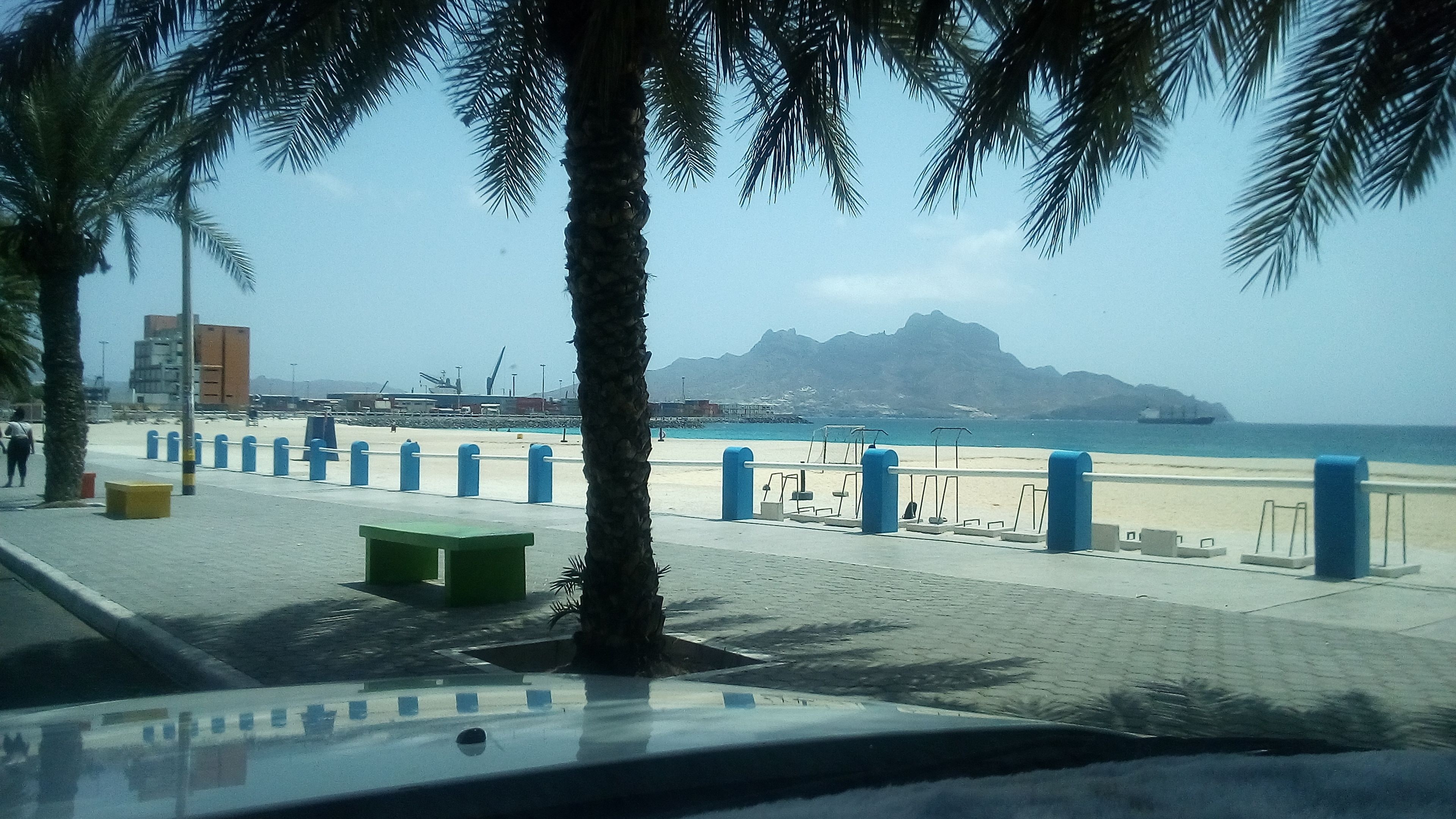 Un paseo marítimo con palmeras, bancos y vistas al mar y a las montañas.