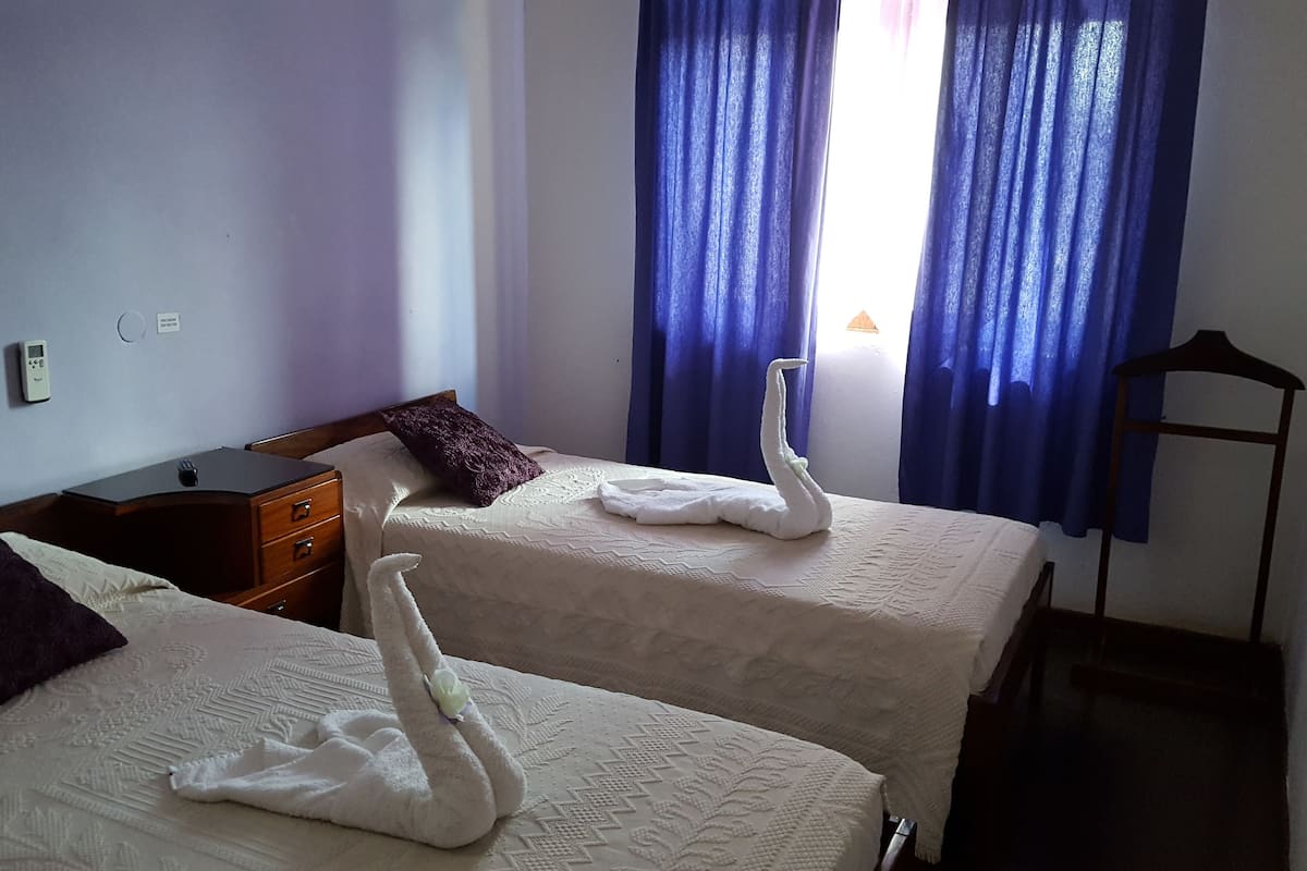 Two beds with white linens, a wooden nightstand, a purple pillow, and a wall-mounted thermostat.