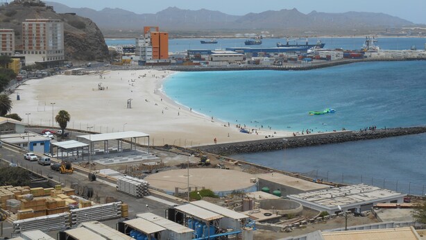 Eine Küstenlandschaft mit Sandstrand, einem Hafen voller Schiffe und Industriegebäuden.