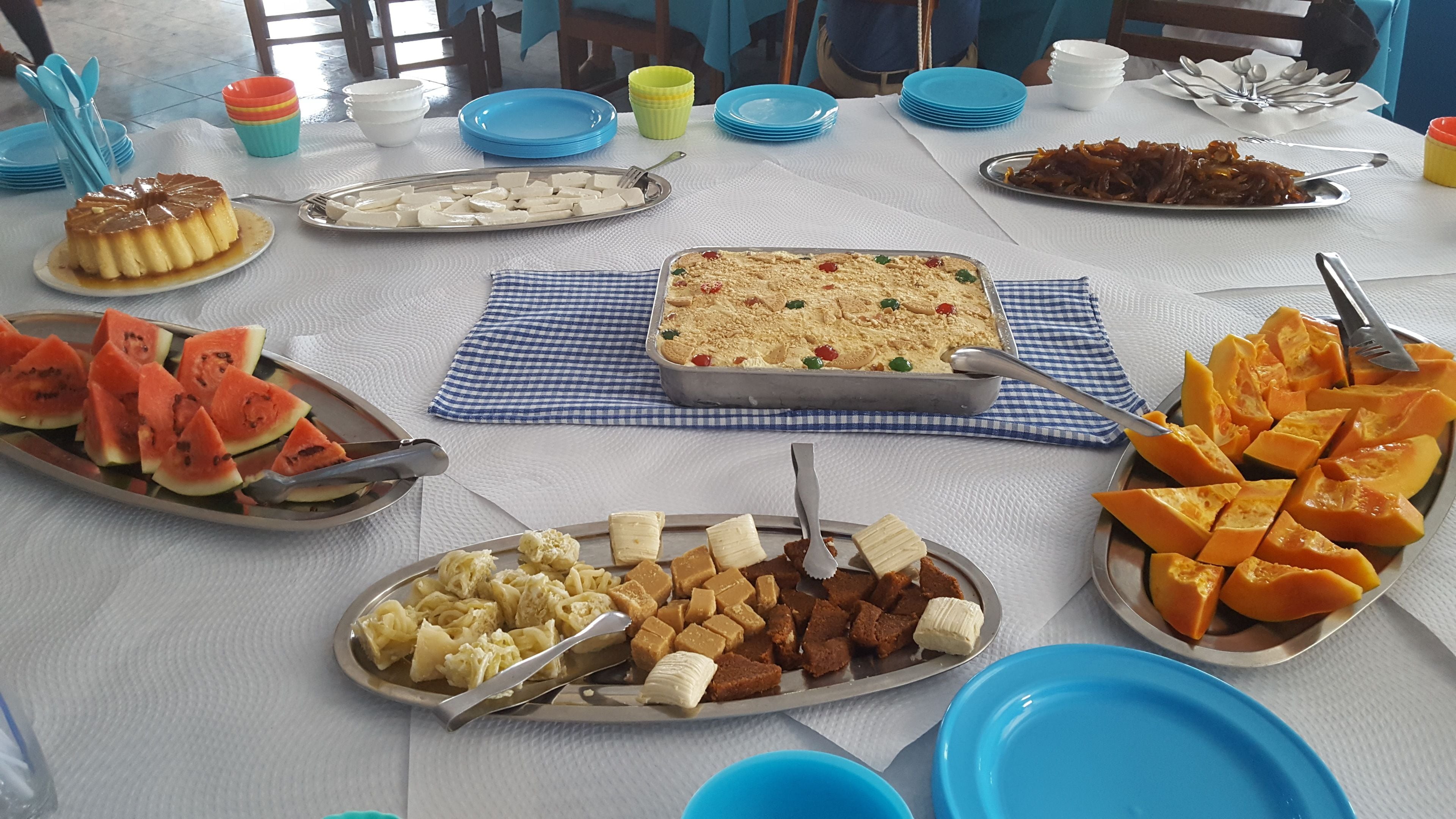 Een tafel gedekt met verschillende gerechten, waaronder een vla, watermeloenpartjes, een ovenschotel en diverse soorten fruit.