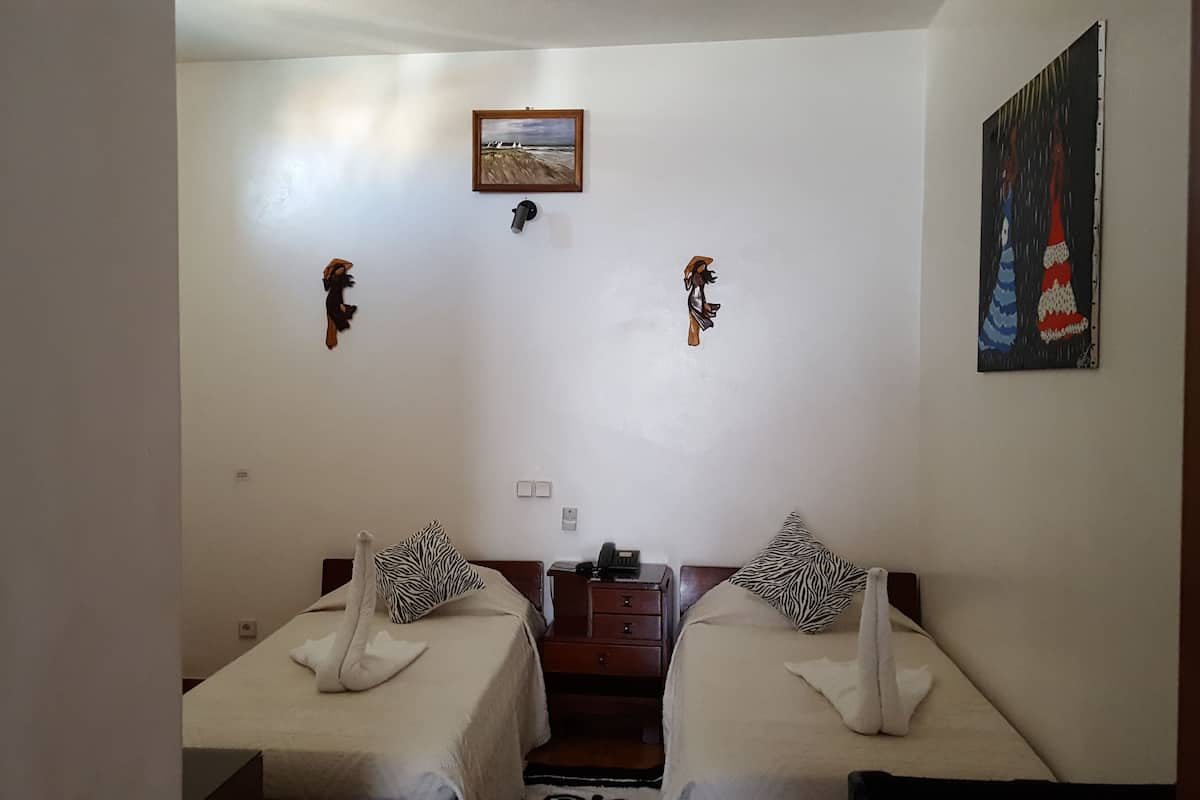 A room with two beds, each with a zebra-patterned pillow and a swan-shaped towel. There is a small table with a telephone and a lamp between the beds. A painting of a beach scene hangs on the wall above the beds.