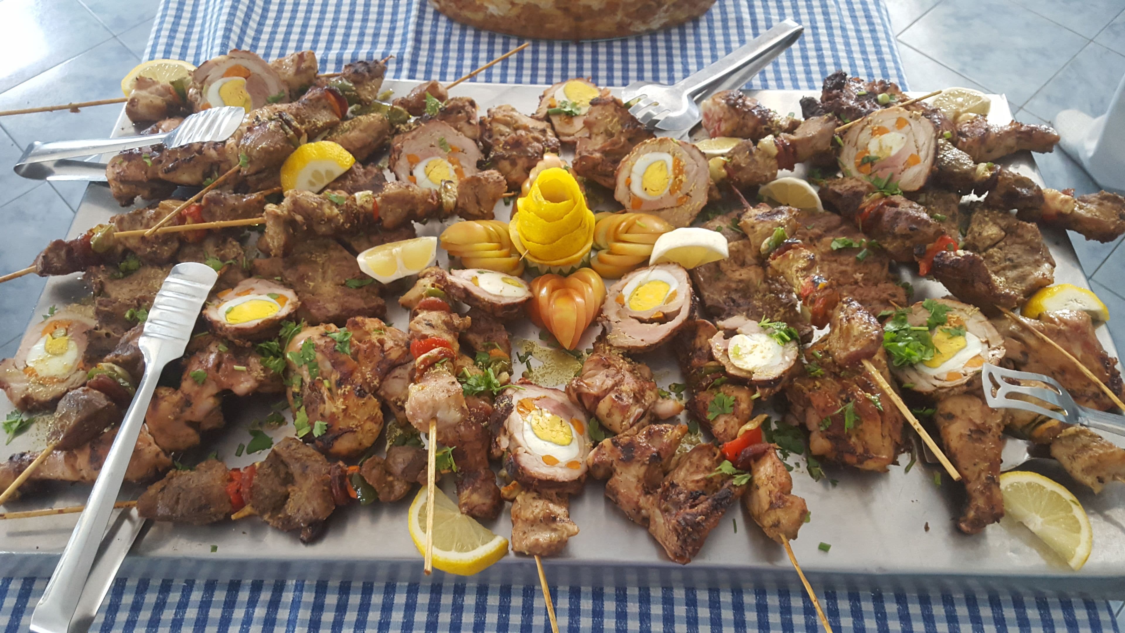 A large platter of skewered meats, some with eggs, garnished with herbs and lemon slices, on a checkered tablecloth.