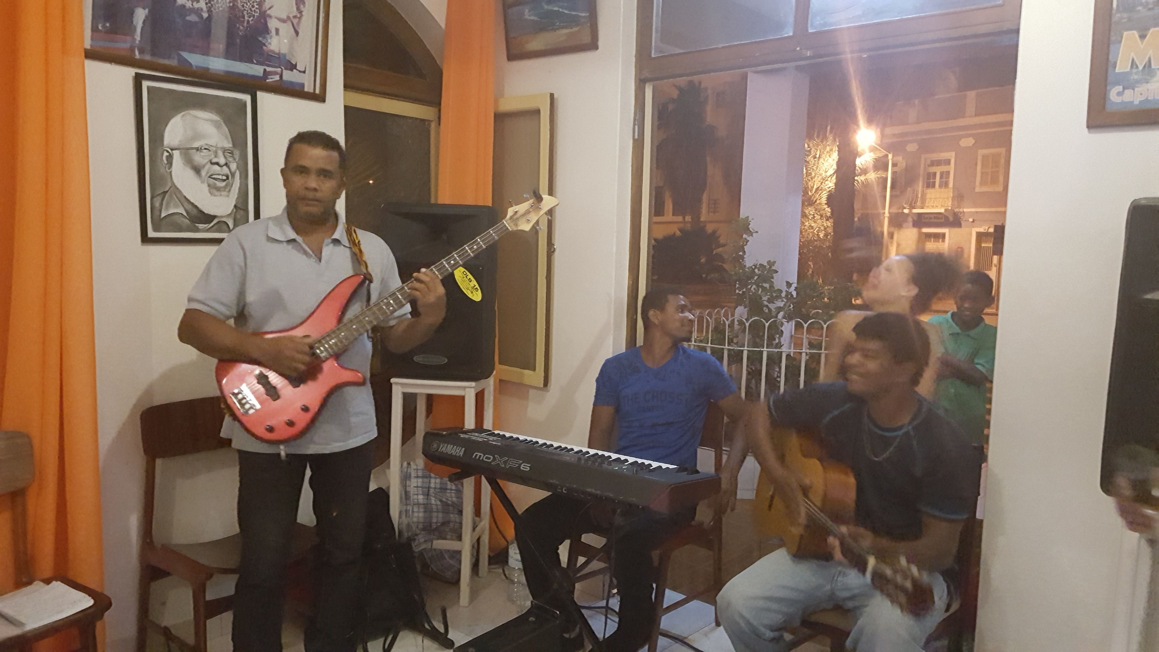 Una habitación con una persona tocando una guitarra eléctrica roja, un teclado Yamaha y otra persona tocando una guitarra acústica. Hay cuadros en la pared y una ventana con vista a edificios.