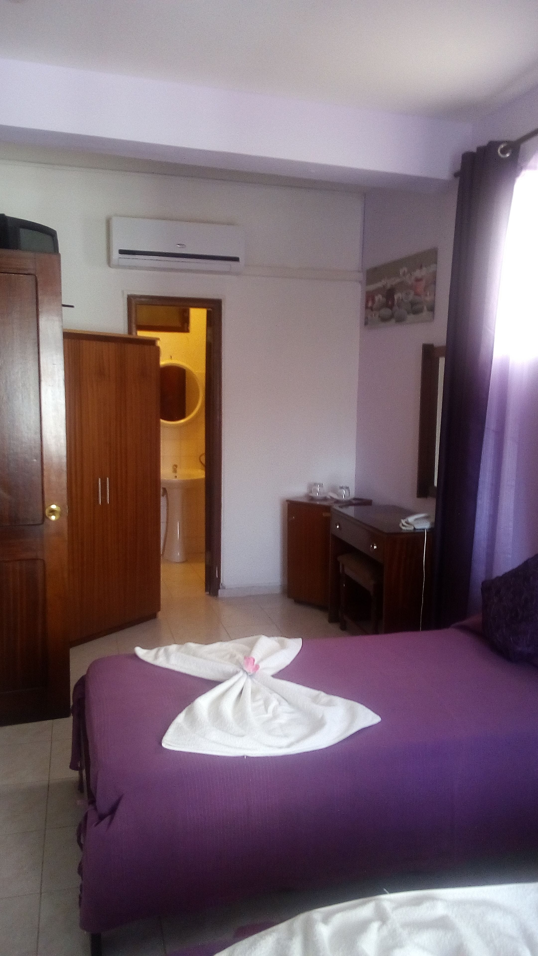 A hotel room with a purple bedspread, a wooden wardrobe, a desk with a chair, an air conditioning unit, and a bathroom visible through an open door.