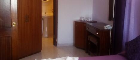 A hotel room with a purple bedspread, a wooden wardrobe, a desk with a chair, an air conditioning unit, and a bathroom visible through an open door.