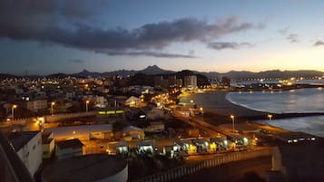 Eine Küstenstadt im Dämmerlicht mit beleuchteten Straßen und Gebäuden, einem Strand und Bergen im Hintergrund.