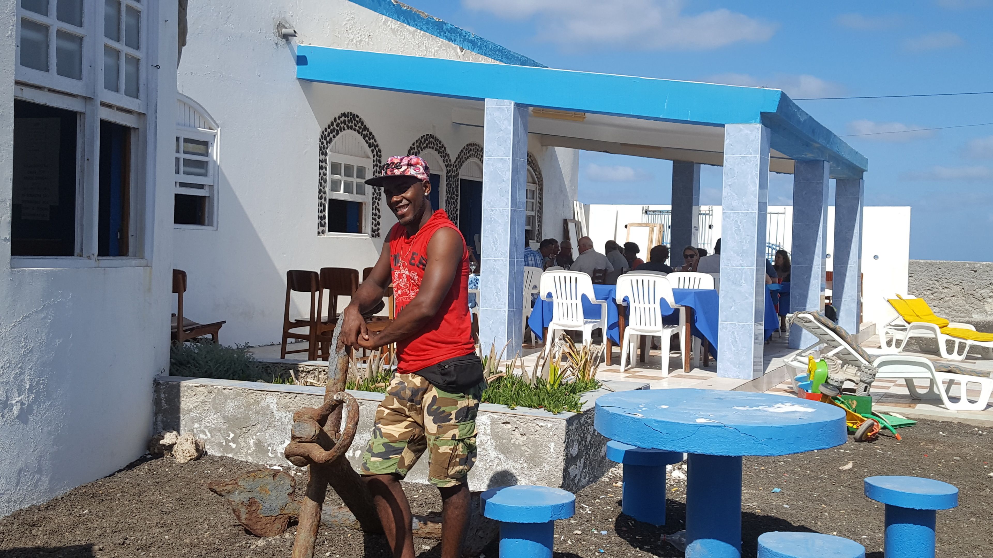 A man standing outside a building with a blue roof and white walls, near a seating area with plastic chairs and tables.