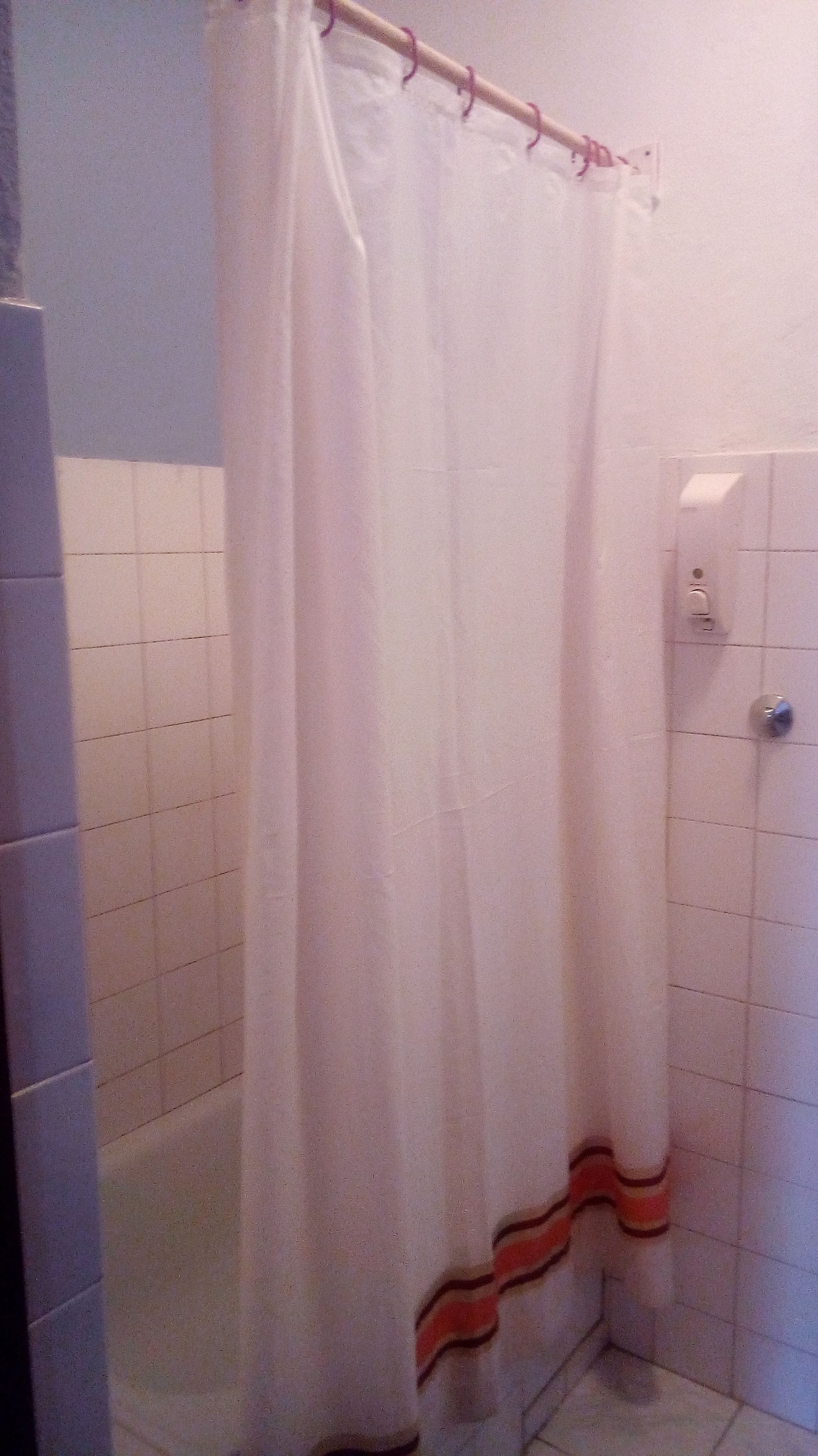 Casa de banho com paredes revestidas a azulejo branco, uma cortina de chuveiro branca com parte inferior listrada em vermelho e branco, e uma porta branca com puxador prateado.