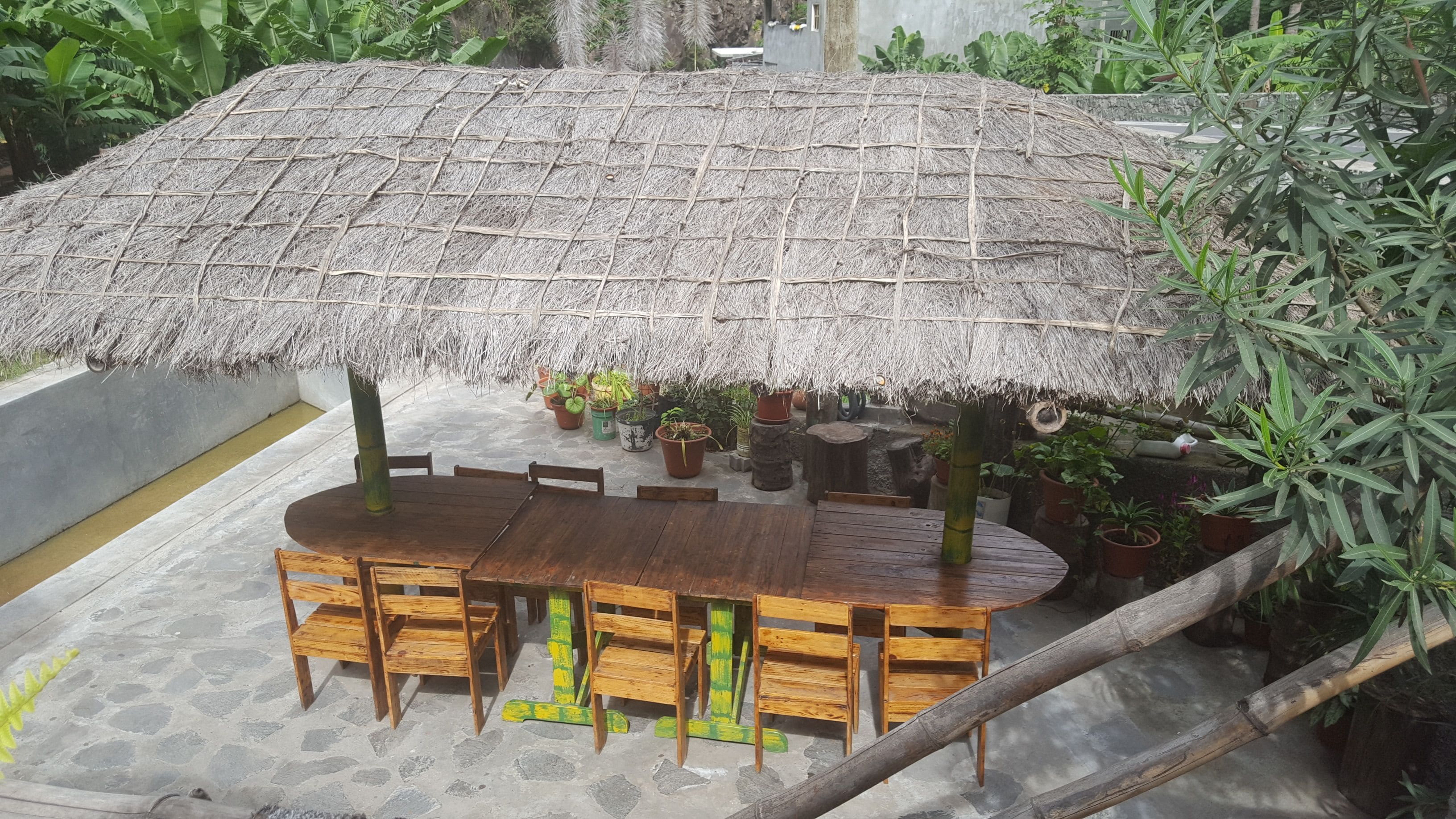 A thatched-roof outdoor dining area with wooden tables and chairs, surrounded by potted plants.