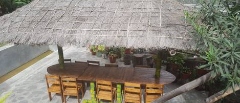A thatched-roof outdoor dining area with wooden tables and chairs, surrounded by potted plants.