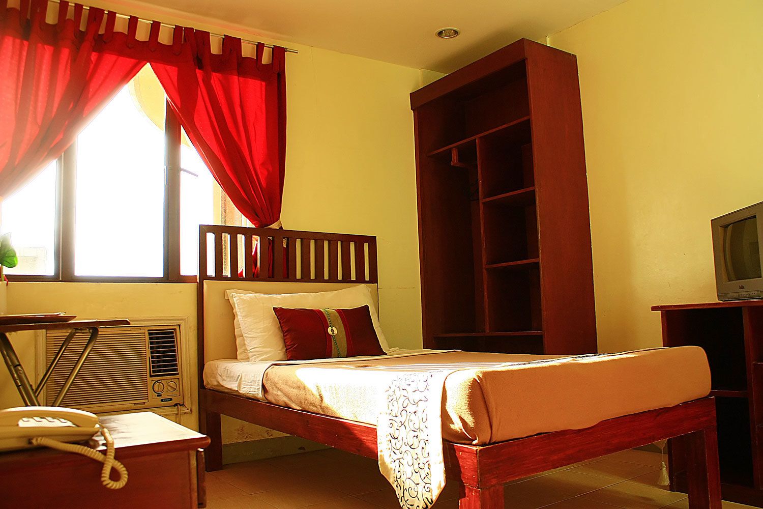 Una habitación con una cama, un escritorio con teléfono, una estantería y un televisor.