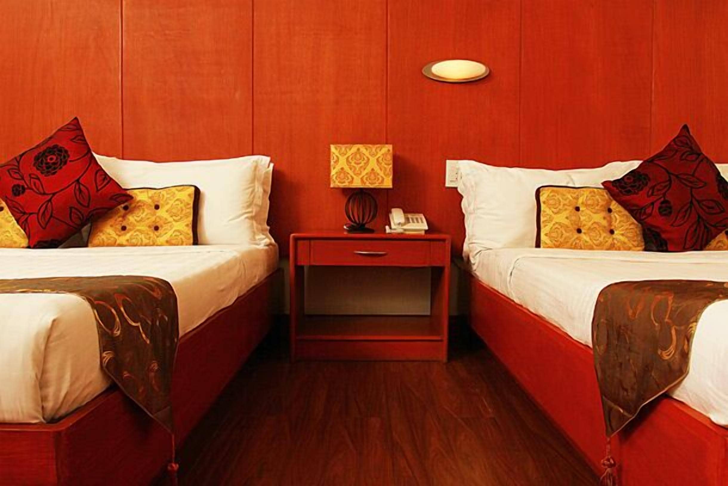 Habitación de hotel con dos camas, cada una con una colcha roja y blanca, una mesita de noche con lámpara y un pequeño cajón.