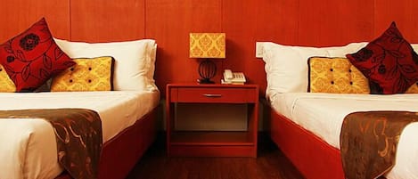 Habitación de hotel con dos camas, cada una con una colcha roja y blanca, una mesita de noche con lámpara y un pequeño cajón.