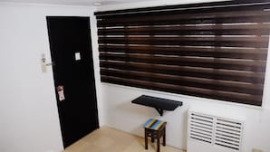 Una habitación con una puerta negra, una ventana con persianas horizontales, un taburete pequeño y un equipo de aire acondicionado blanco.