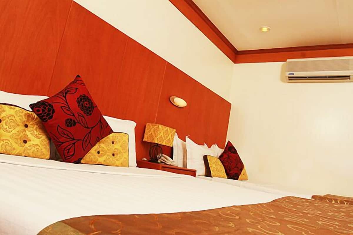 A hotel room with a large bed, multiple pillows, a bedside table with a lamp, and an air conditioning unit on the wall.