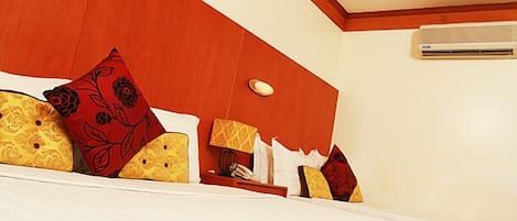 Una habitación de hotel con una cama grande, varias almohadas, una mesita de noche con lámpara y un equipo de aire acondicionado en la pared.