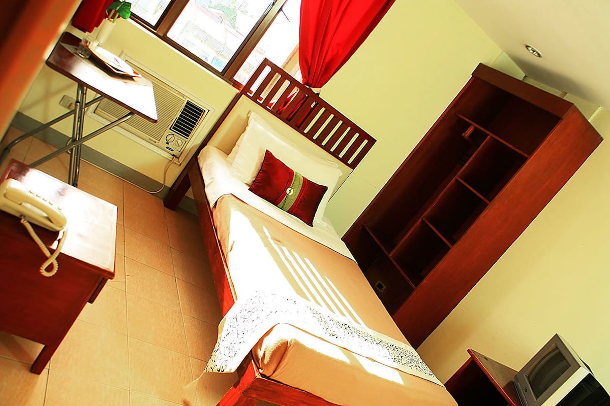 A hotel room with a bed, a desk with a telephone, a window with curtains, and a wardrobe.