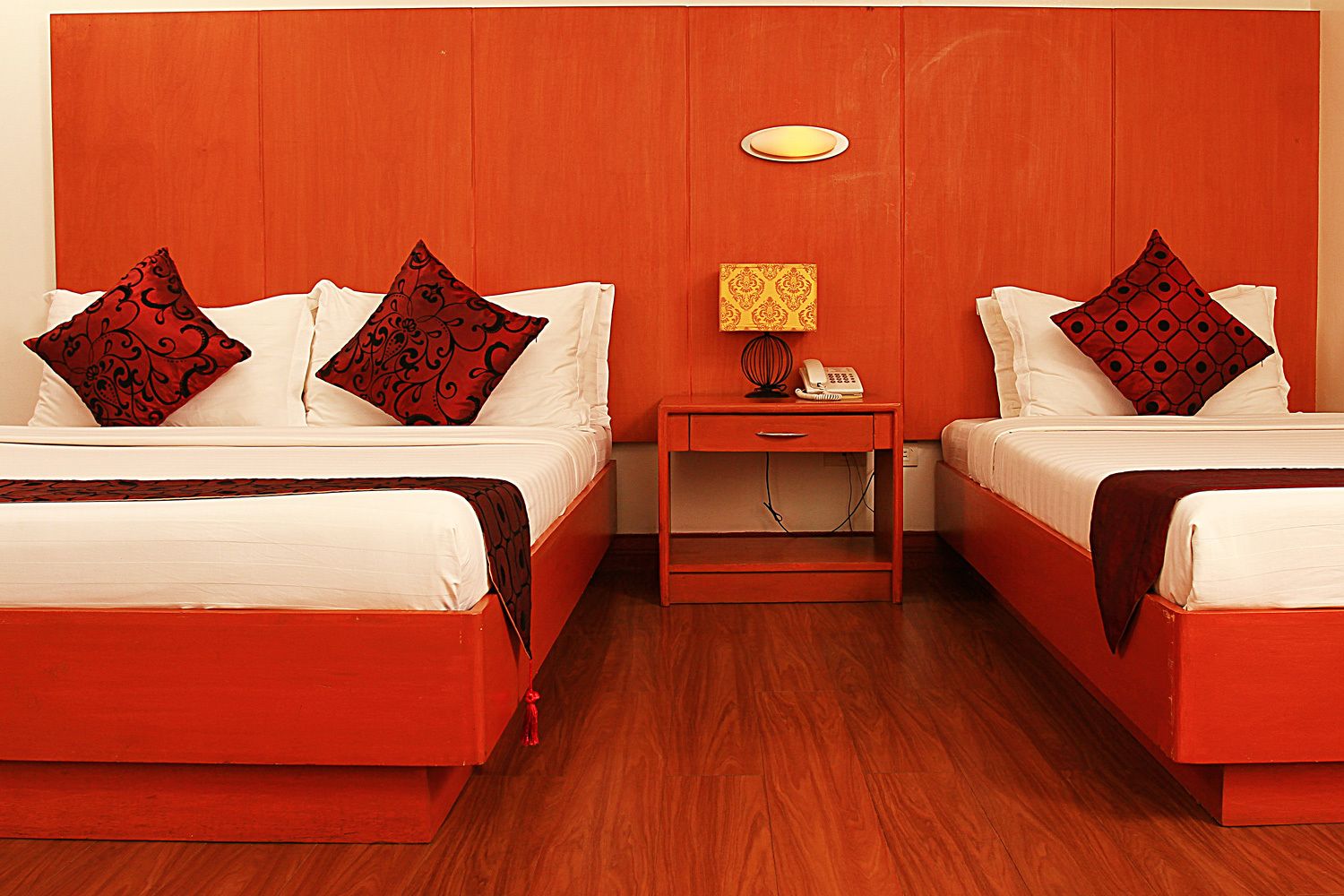 A hotel room with two beds, each with white bedding and red patterned pillows. There is a wooden nightstand with a lamp and a telephone between the beds. The room has a wooden floor and a wooden headboard.