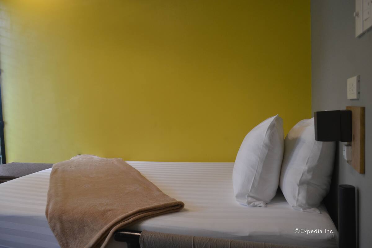 A hotel room with a bed, a brown blanket, white pillows, and a wall-mounted lamp.