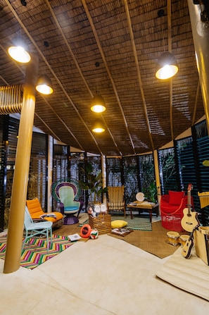 A room with a thatched roof, wooden beams, and a variety of seating including chairs and a sofa. There are musical instruments and a guitar on the floor.