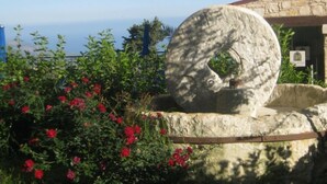 A stone sculpture with a circular hole, surrounded by red flowers and greenery, with a stone building in the background. - Palates Village Hotel (Droushia)