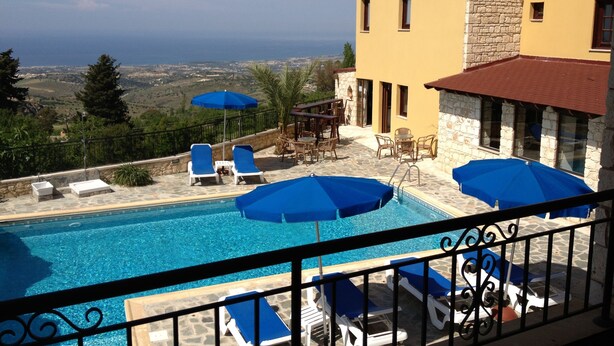 A pool area with blue umbrellas, lounge chairs, and a view of the sea. - Palates Village Hotel (Droushia)