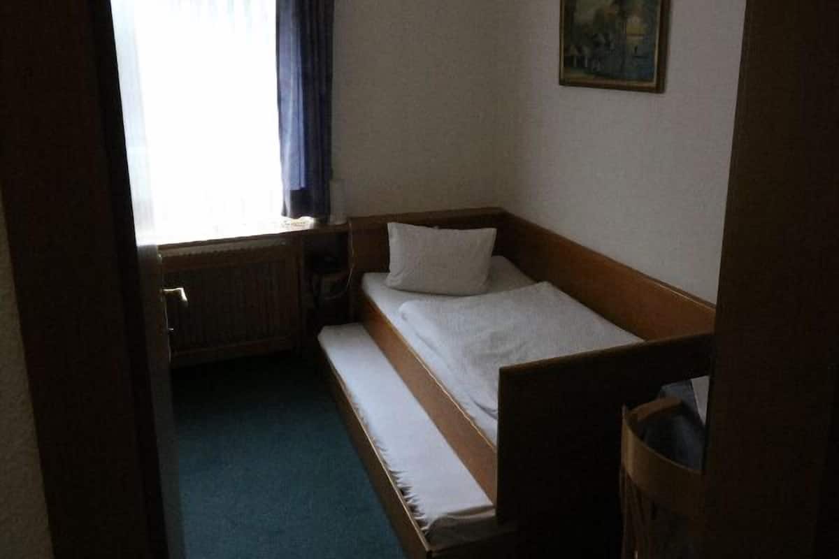 Ein kleines Zimmer mit einem Einzelbett, einem Holzkopfteil, einer weißen Matratze und einem kleinen Nachttisch.