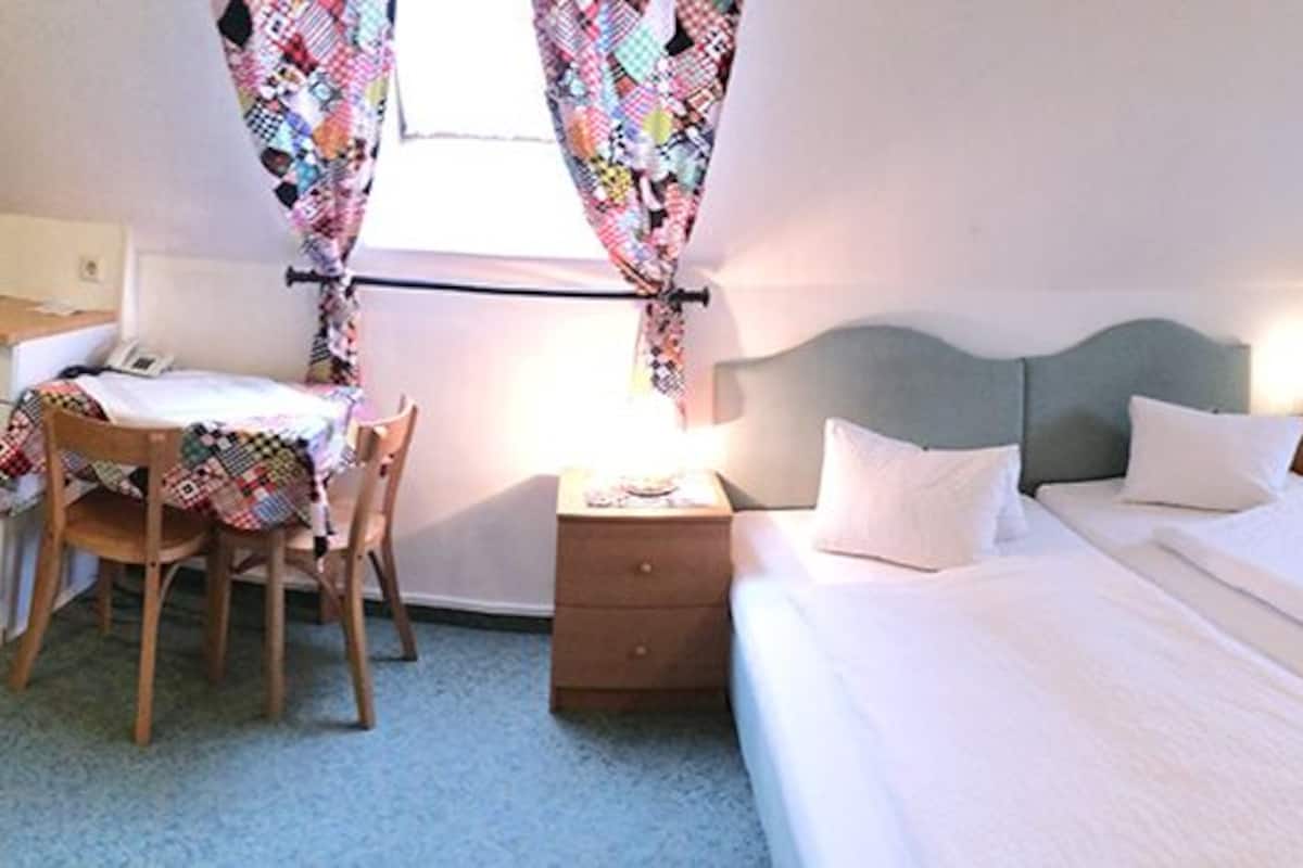 Ein kleiner, kompakter Raum mit einem Einzelbett, einem Schreibtisch mit Stuhl, einer kleinen Küchenzeile und einem Fenster mit Vorhängen.
