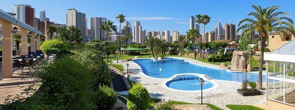A swimming pool with a city skyline in the background, surrounded by greenery and palm trees.