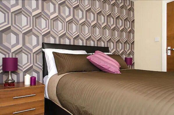 Cama bem arrumada com colcha marrom e várias almofadas, incluindo uma cor-de-rosa, num quarto com papel de parede estampado e uma mesinha de cabeceira de madeira com candeeiro e velas.