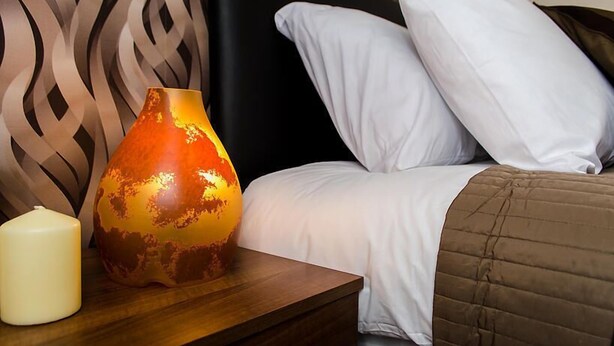 A neatly made bed with white pillows and a brown quilt, a white candle, and a decorative vase on a wooden nightstand. - Trivelles Hotels - Eccles New Road (Salford)
