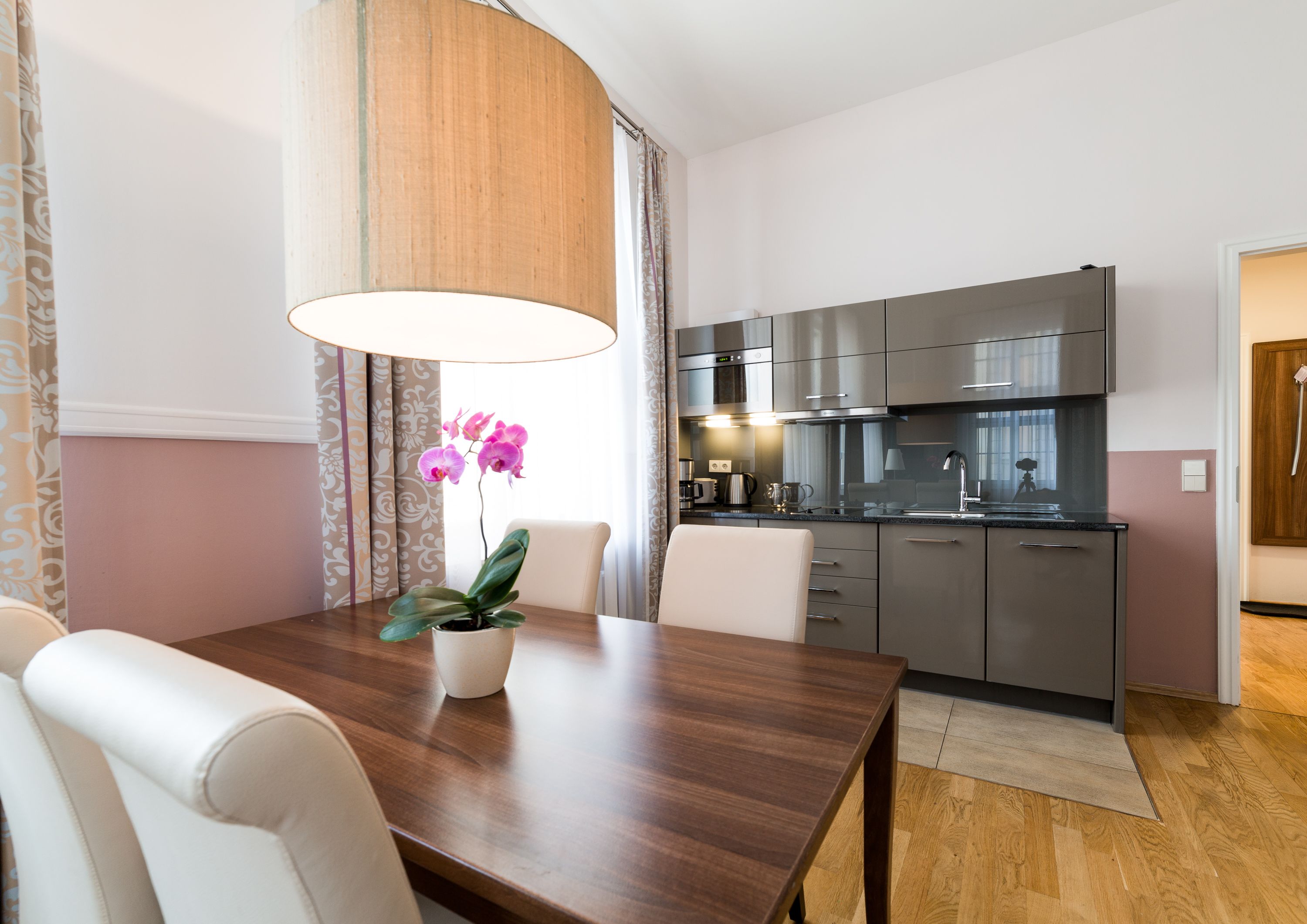 Una cocina moderna con electrodomésticos de acero inoxidable, un comedor con mesa de madera y sillas blancas, y una orquídea en maceta sobre la mesa.