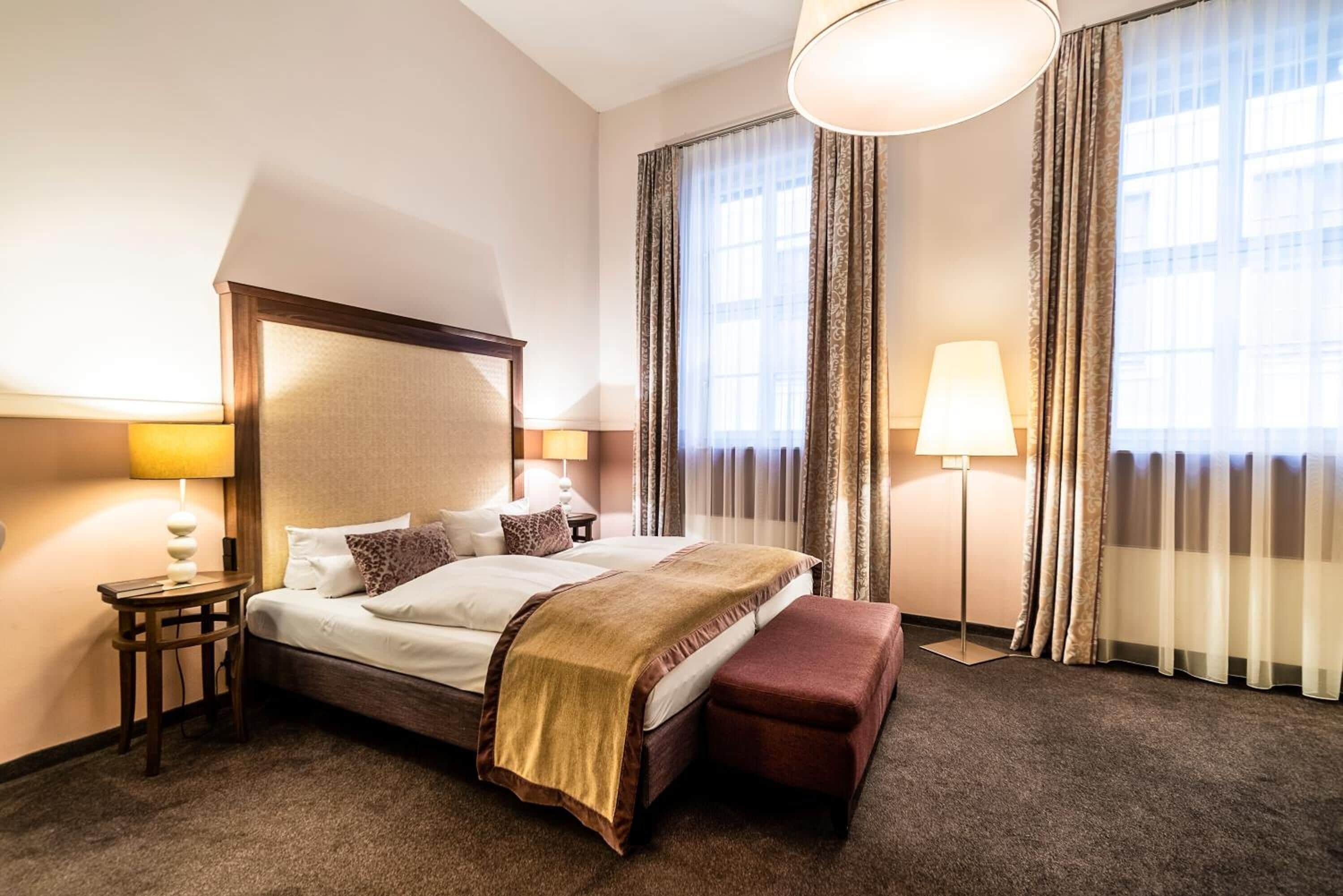 Hotelkamer met een groot bed, een nachtkastje met een lamp, een bankje en een raam met gordijnen.