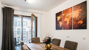 Un comedor con una mesa de madera, seis sillas y un florero con flores. Hay un ventanal con cortinas, y un cuadro de un mapa mundial en la pared.