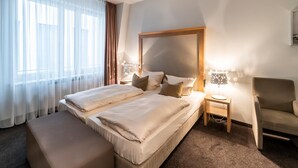 Habitación de hotel con una cama grande, una mesita de noche con lámpara, una silla y una ventana con cortinas traslúcidas.